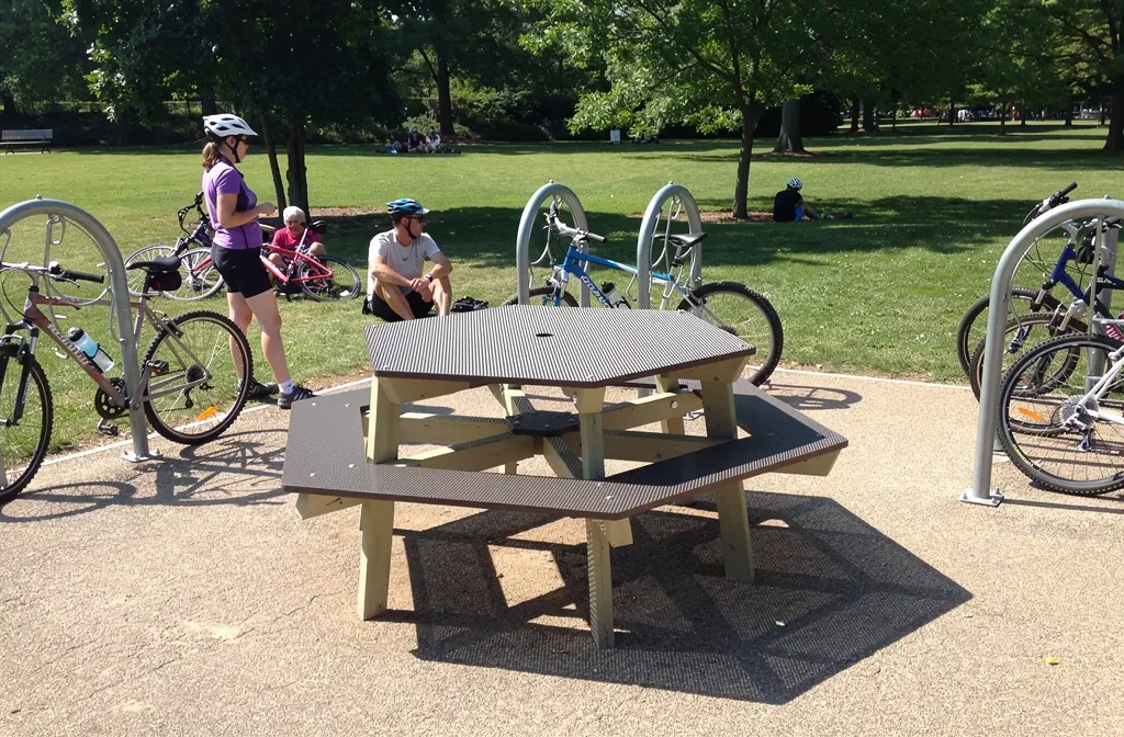 Pièce d’aire de pique-nique en bois avec table et bancs, entourée de vélos attachés à des arceaux métallique à une aire de repos du parc.