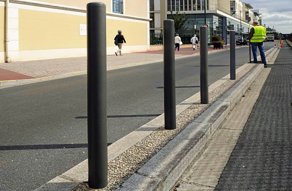 Poteaux de protection en acier à sceller alignés le long d’un trottoir, à proximité d’un parking, avec chaussée et trottoir séparés.